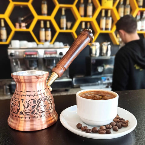 IST Series Thick Solid Copper Engraved Cezve For Stovetop Turkish Greek Arabic Coffee Pot beside full coffee cup in cafe setting