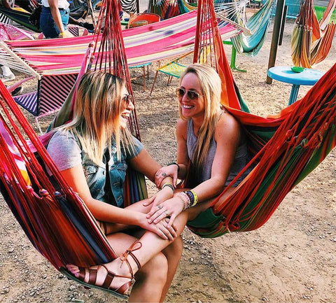 Two women smiling sitting in colorful hammock chairs outdoors; image for Hammock Chair Hanging Rope Swing Seat For Indoor And Outdoor Use Sturdy Cotton Weave Cozy Chill Lounge Swing Supports Up To 300 Lbs Easy Hanging Setup Gray.