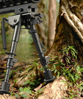 6 To 9 Inch Picatinny Bipod With Adjustable Five Setting Legs And Spring Return Quick Attach Picatinny Adapter Included Heavy Duty Steel And Aluminum Construction Black Anodized Finish For Rugged Hunting And Shooting among forest greenery.