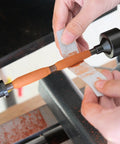 Pen mandrel MT2 on woodturning lathe; orange wooden pen blank being sanded with sandpaper wrap.