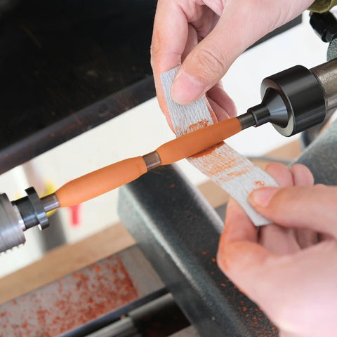 Pen mandrel MT2 on woodturning lathe; orange wooden pen blank being sanded with sandpaper wrap.