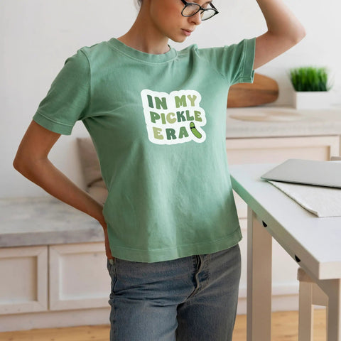 Woman wearing green shirt with "In My Pickle Era" text, next to iron-on heat transfer paper for inkjet printers, suitable for dark and light fabrics.
