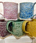 Five colorful embossed ceramic mugs in pink, blue, purple, green, and yellow, stacked in a kiln, showcasing intricate designs.
