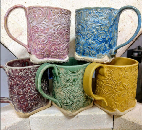 Five colorful embossed ceramic mugs in pink, blue, purple, green, and yellow, stacked in a kiln, showcasing intricate designs.
