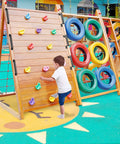 Child climbing a wooden frame with colorful grips, next to a tire-based climbing structure on an outdoor playground.