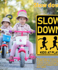 Two children on tricycles next to a Slow Down Kids At Play Road Sign For Neighborhoods, Aluminum Weatherproof Safety Plaque.