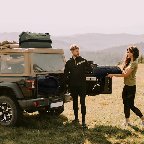 Couple loading Waterproof Duffel Backpack 60L Heavy Duty Dry Gear Bag For Camping Boating And Outdoor Travel into SUV in a field.