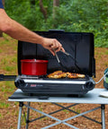 Person using 2-In-1 Tabletop Camping Grill And Stove With Two Burners, 20,000 BTUs Of Cooking Power, cooking chicken outdoors.