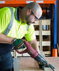 Worker using a power tool on wood wearing high-visibility shirt and safety glasses in warehouse