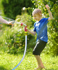 Garden hose, blue with red spray nozzle, held by child while spraying water in a sunlit backyard.