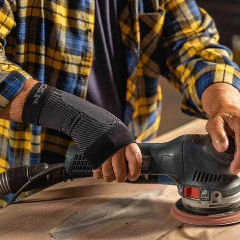 Person using a power sander while wearing WS6 Compression Wrist Sleeve for Carpal Tunnel Relief and All-Day Pain Support, Reducing Strain and Fatigue with K-Zone Therapy For Everyday Comfort, Lightweight Support, And Steady Wrist Stabilization in black.