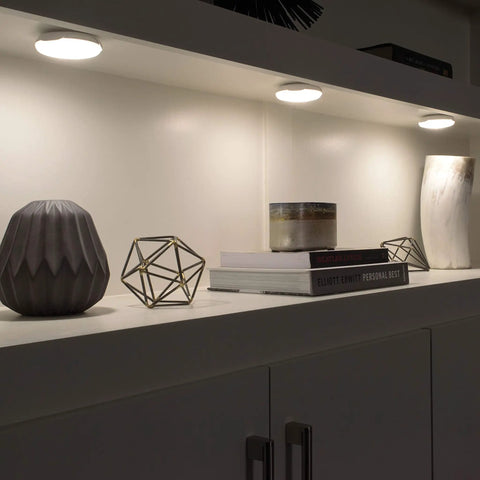 Battery Operated LED Puck Light For Under Cabinets And Shelves With Motion Sensor And Auto Shut Off After 20 Seconds 30 Lumen Bright White Cabinet Lighting No Fuss Easy Install illuminating a shelf with decorative items