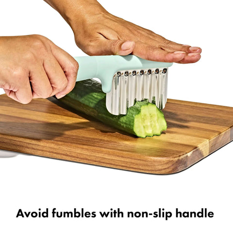OXO Tot crinkle cutter for baby feeding, mint green handle with stainless steel blades cutting cucumber on a board