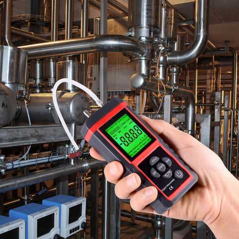 Close-up of red and black digital manometer held in hand with green LCD display, connected to HVAC piping.