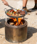 Stainless Steel Grill Cookware For Solo Stove Bonfire 19.5 Inch Fire Pit Portable Round Grilling Pan With Legs For Outdoor BBQ Camping And Backyard Cooking Adventures being used to grill food over an open flame.