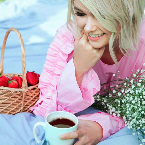 Woman in pink dress holding a coffee cup with a picnic basket of strawberries on a blue blanket outdoors.