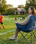 Woman sitting on heavy duty portable camping chair with 350 lb capacity next to a soccer field, focus on ergonomic padded seat.