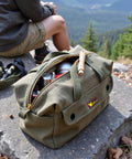 Tough As Nails Waxed Canvas Military Duffle Bag with Water Resistance, Adjustable Strap, on a rocky outdoor setting.