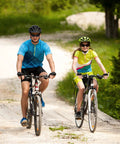 Two people biking on a rural path wearing helmets and colorful cycling gear, riding mountain bikes on a sunny day.