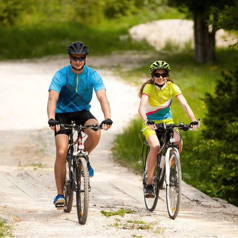 Two people biking on a rural path wearing helmets and colorful cycling gear, riding mountain bikes on a sunny day.