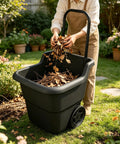 Person using Heavy Duty 15 Gallon Rolling Garden Cart With Retractable Handle And 7 Inch Wheels For Outdoor Yard Work A Real Workhorse Lightweight Resin Utility Wagon For Gardening Lawn And Home Hauling, placing dry leaves into black durable cart on grass with garden in background