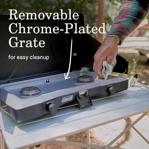 Triton Two Burner Propane Camp Stove With Wind Guards being cleaned, showing removable chrome-plated grate on cooktop