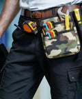 Person wearing tool belt with camouflage pouch holding various hand tools like wrench, pliers, and measuring tape.
