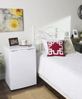 White mini fridge next to a bed with a red pillow, in a decorated bedroom with a wall clock and floral artwork.