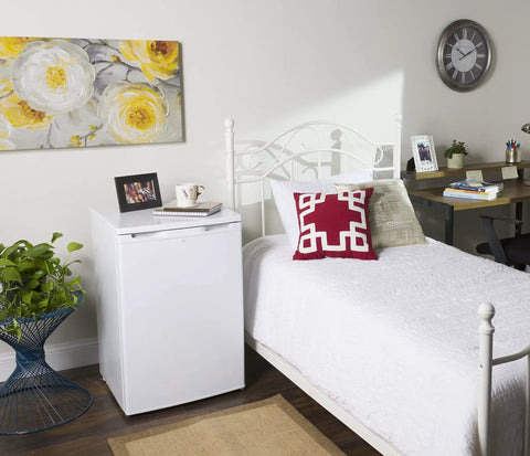 White mini fridge next to a bed with a red pillow, in a decorated bedroom with a wall clock and floral artwork.