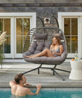 Double Egg Swing Chair With Stand For Outdoor Patio Hand Made Rattan Wicker Two Hanging Egg Chairs Hammock Chair With UV Resistant Cushions And Solid Metal Frame, Porch Loveseat For Backyard Chill shown by a poolside with a woman seated and a man in the pool.