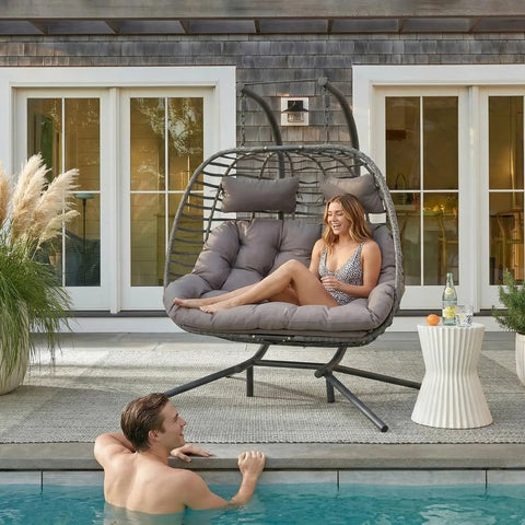 Double Egg Swing Chair With Stand For Outdoor Patio Hand Made Rattan Wicker Two Hanging Egg Chairs Hammock Chair With UV Resistant Cushions And Solid Metal Frame, Porch Loveseat For Backyard Chill shown by a poolside with a woman seated and a man in the pool.