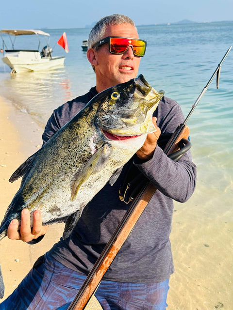 Person holding large fish on beach wearing sunglasses and holding a fishing rod, with a boat in the background.