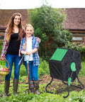 Compost Tumbler Bin With Sliding Doors, 18.5 Gallon Rotating Chamber in garden setting with two people, ideal for kitchen and yard waste.