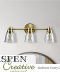 Three-light metal bathroom vanity wall light fixture in brass with clear glass shades mounted on a wall above a mirror.