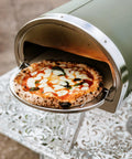 Pizza being cooked in green Gozney Roccbox, showcasing its outdoor use.
