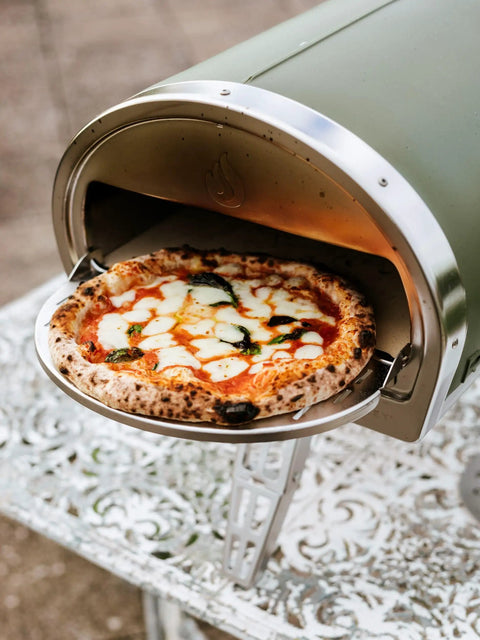 Pizza being cooked in green Gozney Roccbox, showcasing its outdoor use.