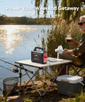 Go To Portable Solar Generator 296Wh Kit on table by lake, man holding cup, power station and fan visible, outdoors setup