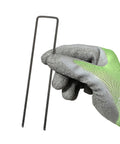 Gloved hand holding 100 Pack 6 Inch 11 Gauge Heavy Duty U Shaped Garden Staples Ground Stakes For Anchoring Landscape Fabric Spikes Pins Pegs