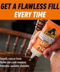 All Purpose Wood Filler Natural Color 6 Ounce Tube Pack Of 1 held by a hand, highlighting smooth table repair, indoors.