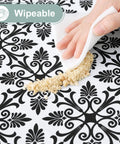 Hand wiping crumbs on black patterned Snug Fit Elastic Fitted Vinyl Table Cover For 6 Foot Tables, Non Slip Waterproof Flannel Backed Plastic Tablecloth, Stain Proof, Ideal For Picnics, Camping, Outdoor Events, Black 30x72 Inches.
