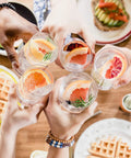Group toasting with fizzy drinks and fruits over brunch table with waffles and avocado toast.
