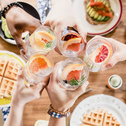 Five people toasting with sparkling drinks in glasses over a brunch table with waffles and avocado toast visible.