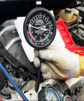Front view of a black HFS fuel pump tester gauge with white dial, held by a gloved hand under the hood.
