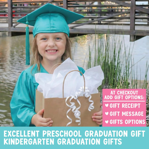Child in graduation cap holding gift bag for preschool graduation near a pond, smiling.