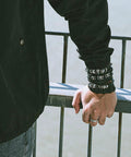 Badass Six Piece Braided Leather Bracelet Set For Men And Women Adjustable Hemp Rope Wristbands With Classic Skull Charm Punk Rock Style Fresh Daily Bling worn by a person resting on a railing.