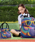 Child sitting with colorful hippo-themed backpack and lunch bag on grass.