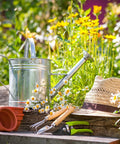 Garden tools with stainless steel root rake, three-prong design, non-slip handle, surrounded by flowers and a straw hat.