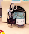 Office jars labeled with black and white dry erase tape, holding office supplies like scissors and tacks, on a desk.