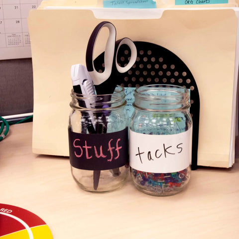 Office jars labeled with black and white dry erase tape, holding office supplies like scissors and tacks, on a desk.