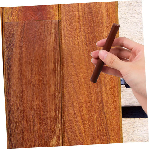 Hand holding dark brown wax filler stick from Dark Brown Wood Repair Kit Wax Filler Sticks For Floors Cabinets And Surfaces Easy To Use No Mess Scratch Repair Crayons That Hide Scuffs And Scratches Fast Fix For Furniture, near wooden surface.